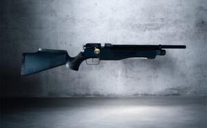 Side view of a shotgun resting on the floor against a textured gray wall.